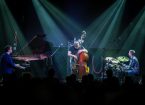 A group of musicians giving a concert on stage, including a piano player, drummer, and stand-up bass.