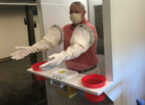 Photo of a man in an enclosed booth with his hands and arms encased in protective gloves. There is a shelf capable of holding supplies connected to the outside of the booth.